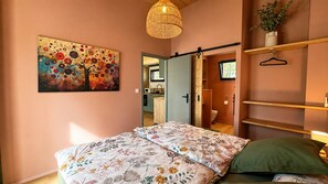 Ein Schlafzimmer mit einem Bett, einem Gemälde, einer Pendelleuchte und Blick in die Küche.