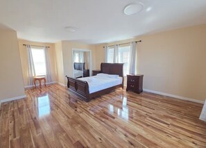 Quarto com piso de madeira, cama, mesa de cabeceira, cômoda e janela com cortinas.