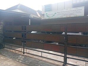 Uma área cercada com pallets de madeira empilhados, um portão metálico e um edifício com uma placa de sinalização ao fundo.