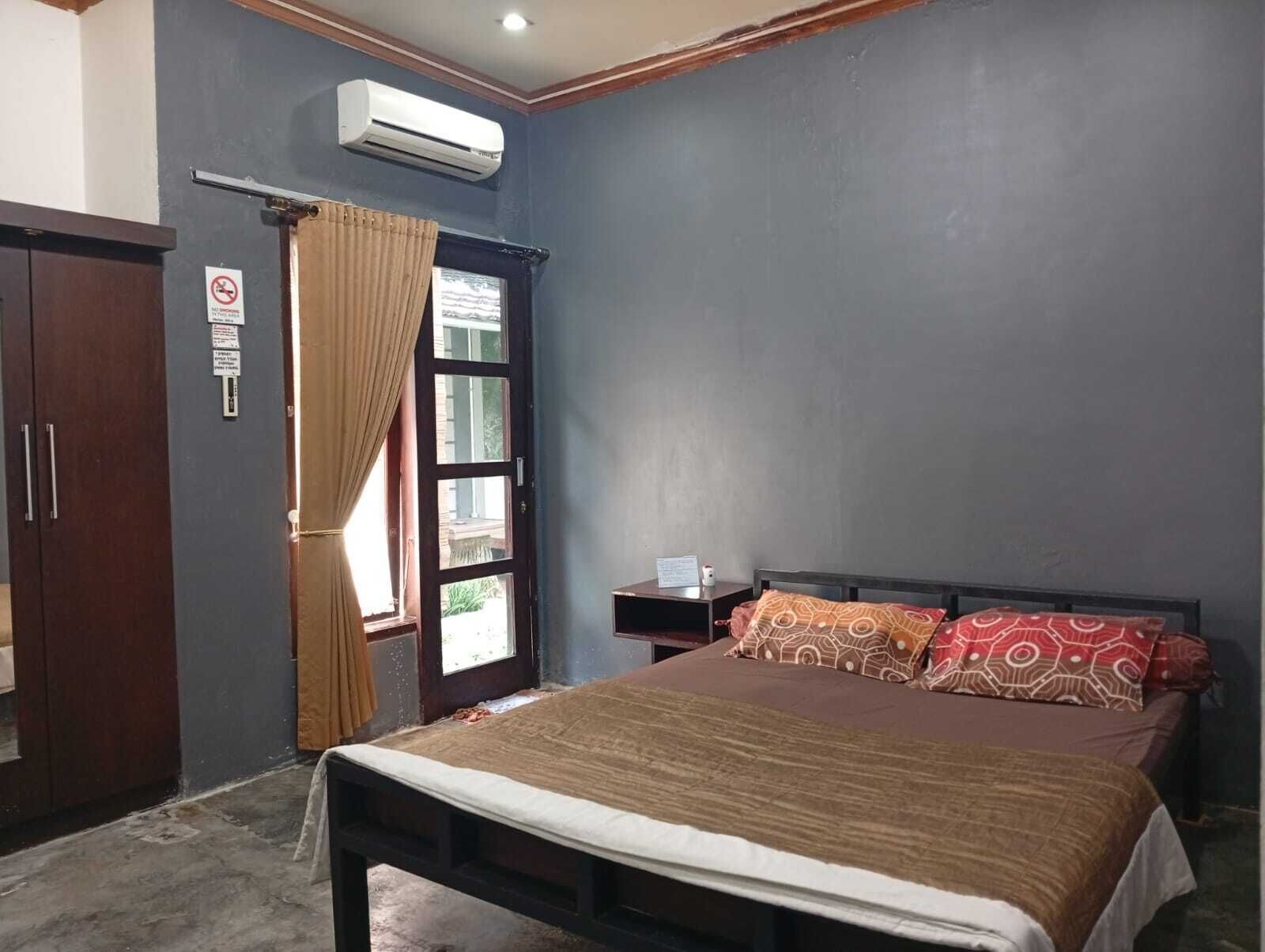 Quarto com cama, mesa de cabeceira, guarda-roupa, ar-condicionado e janela com cortinas.