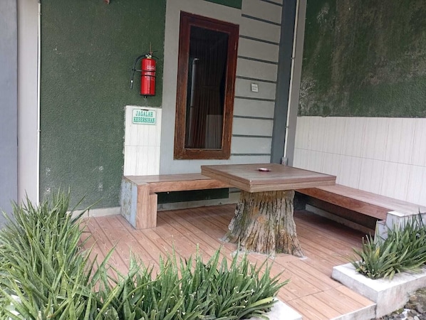 Un ensemble de banc et de table en bois, posĂ© sur une terrasse en bois dont le piĂ©destal est constituĂ© dâune souche dâarbre, le tout entourĂ© de plantes vertes.