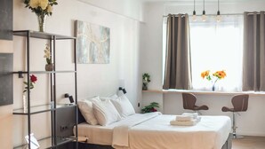 A modern bedroom with a bed, bedside tables, a window with curtains, and a wall-mounted shelf with flowers. - Stylish Studio KingBed&Workspace-WkndNse (Athina)