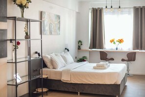 A modern bedroom with a bed, bedside tables, a window with curtains, and a wall-mounted shelf with flowers. - Stylish Studio KingBed&Workspace-WkndNse (Athina)