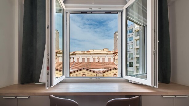 A room with a large window, two chairs, and a view of a building with red-tiled roofs. - Stylish Studio KingBed&Workspace-WkndNse (Athina)