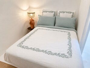 Een net opgemaakt bed met een witte spreideken met een groen bladpatroon, twee kussens, een bedlampje en een klein nachtkastje van hout.