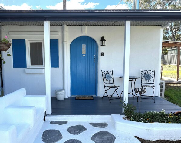 Ett hus med en blå dörr, vita väggar och en veranda med två stolar och ett litet bord.