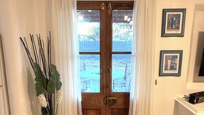 Una stanza con una porta in legno, tende bianche, un vaso con foglie verdi, una sedia in vimini e quadri appesi al muro.
