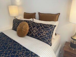Een net opgemaakt bed met gestreepte lakens en decoratieve kussens, een bedlampje en een houten nachtkastje.