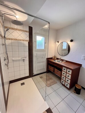 Une salle de bain avec une douche à parois de verre, un carrelage blanc au sol, un meuble-lavabo en bois avec des tiroirs, un miroir et une fenêtre.