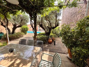 Un patio con una mesa de vidrio, sillas de metal, plantas en macetas y vista a una piscina.