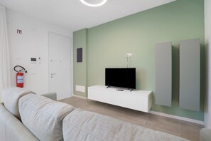 A modern living room with a sofa, a flat-screen TV on a white cabinet, a fire extinguisher, and a door. - Apartment 'Villa Luna 9' with Private Terrace, Balcony and Air Conditioning (Lido di Jesolo)