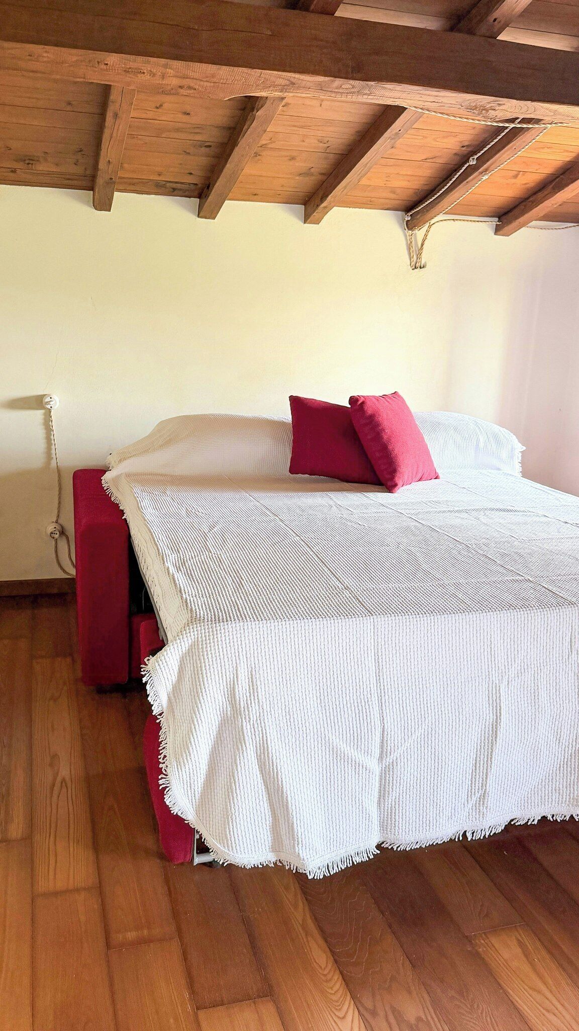 A bedroom with a wooden ceiling, a bed with a white bedspread and red pillows, a red chair, and a white lamp.