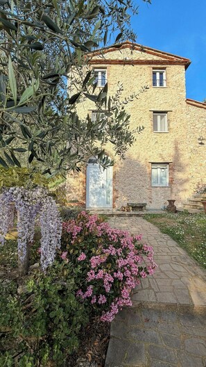 Un bâtiment en pierre doté d’une porte centrale et de fenêtres, entouré d’un jardin où poussent des fleurs violettes et roses.