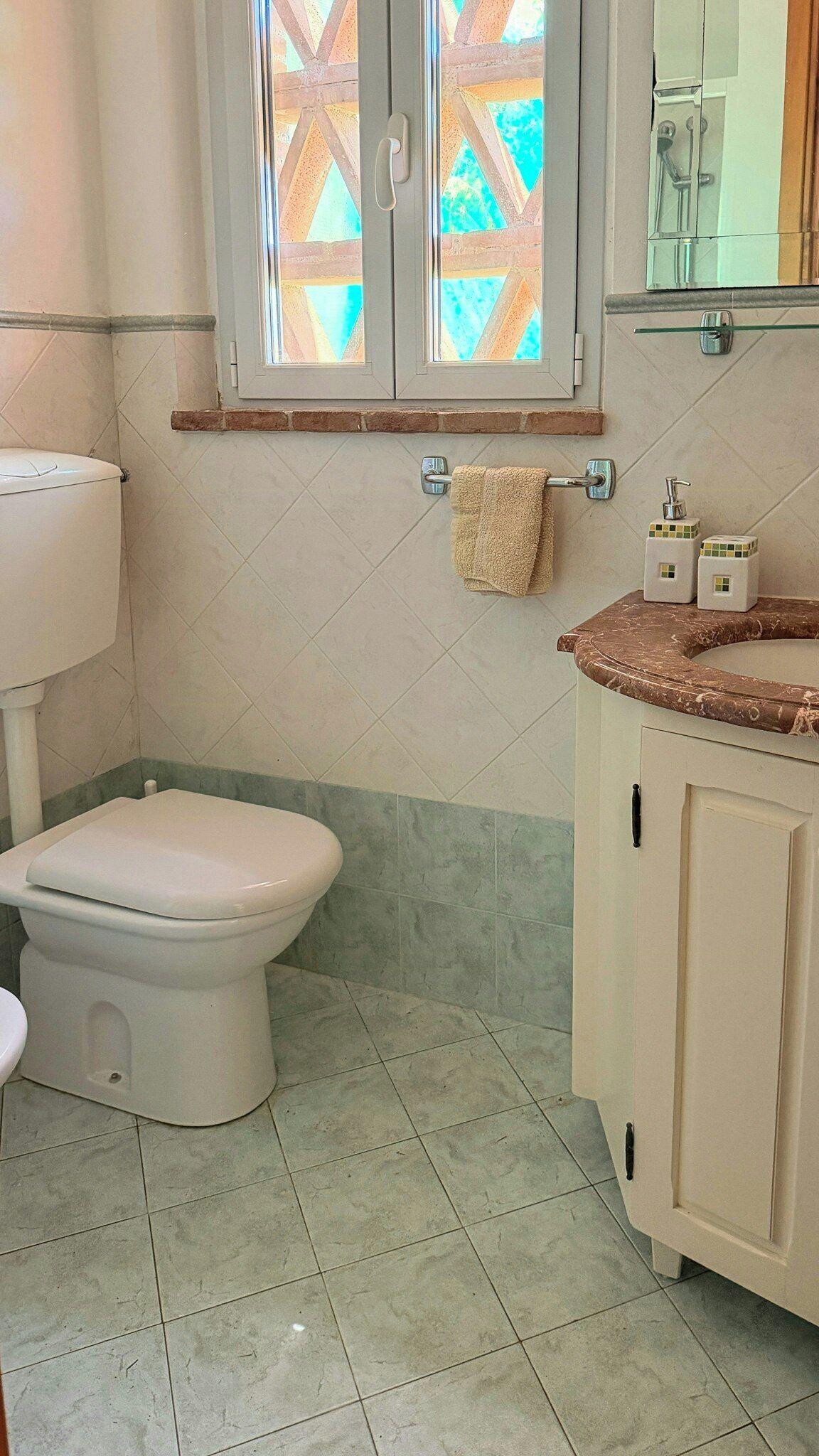 A bathroom with a toilet, a sink with a marble countertop, and a window with a geometric pattern.
