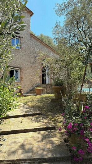 Un bâtiment en pierre doté d’une fenêtre, d’un escalier menant à une porte et entouré de verdure.