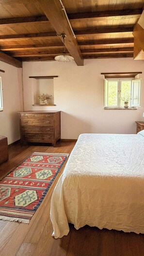 Une chambre avec un plafond en bois, un lit recouvert d’une courtepointe blanche, une commode en bois, un tapis et une fenêtre laissant entrer la lumière du jour.