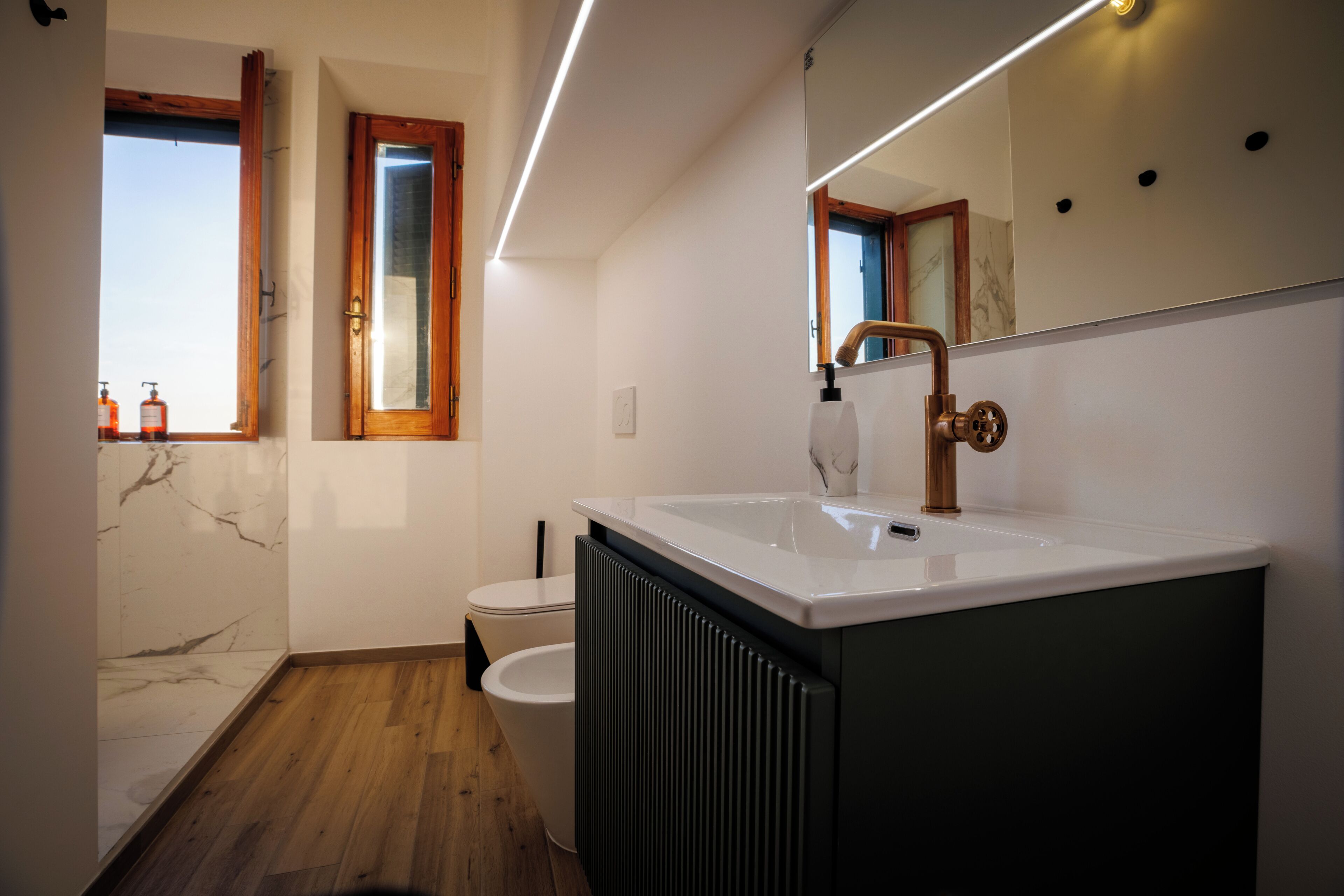 Un baño moderno con un lavamanos blanco, un espejo, un inodoro y una ventana con marcos de madera.
