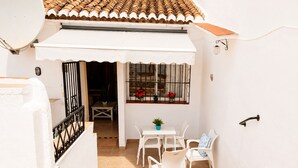 Un bâtiment blanc avec un toit recouvert de tuiles, une antenne parabolique, une petite table avec des chaises et une fenêtre ornée de barreaux en métal et de fleurs rouges.
