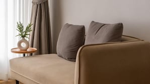Un sofá moderno con dos cojines, una mesita redonda con una planta y una ventana con cortinas.