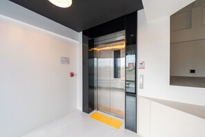 Ein moderner Aufzug mit Glastür, gelber taktiler Bodenbelag am Eingang und einem Feuerschutzmelder an der Wand.