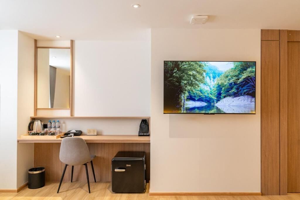 A modern hotel room with a wooden desk, a grey chair, a black storage unit, a trash bin, and a mounted TV displaying a forest scene.