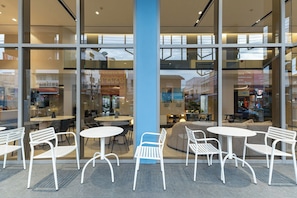 Un espacio interior moderno con asientos, sillas y mesas de metal blanco, ventanas amplias y un pilar azul.