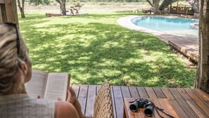 Una persona leyendo un libro en una terraza de madera con una piscina y follaje al fondo.