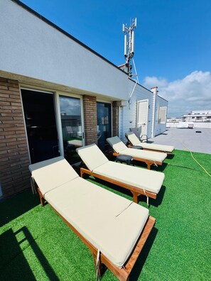 Eine Dachterrasse mit vier Liegestühlen, einer Ziegelwand, einer Schiebeglastür und einem weißen Gebäude im Hintergrund.