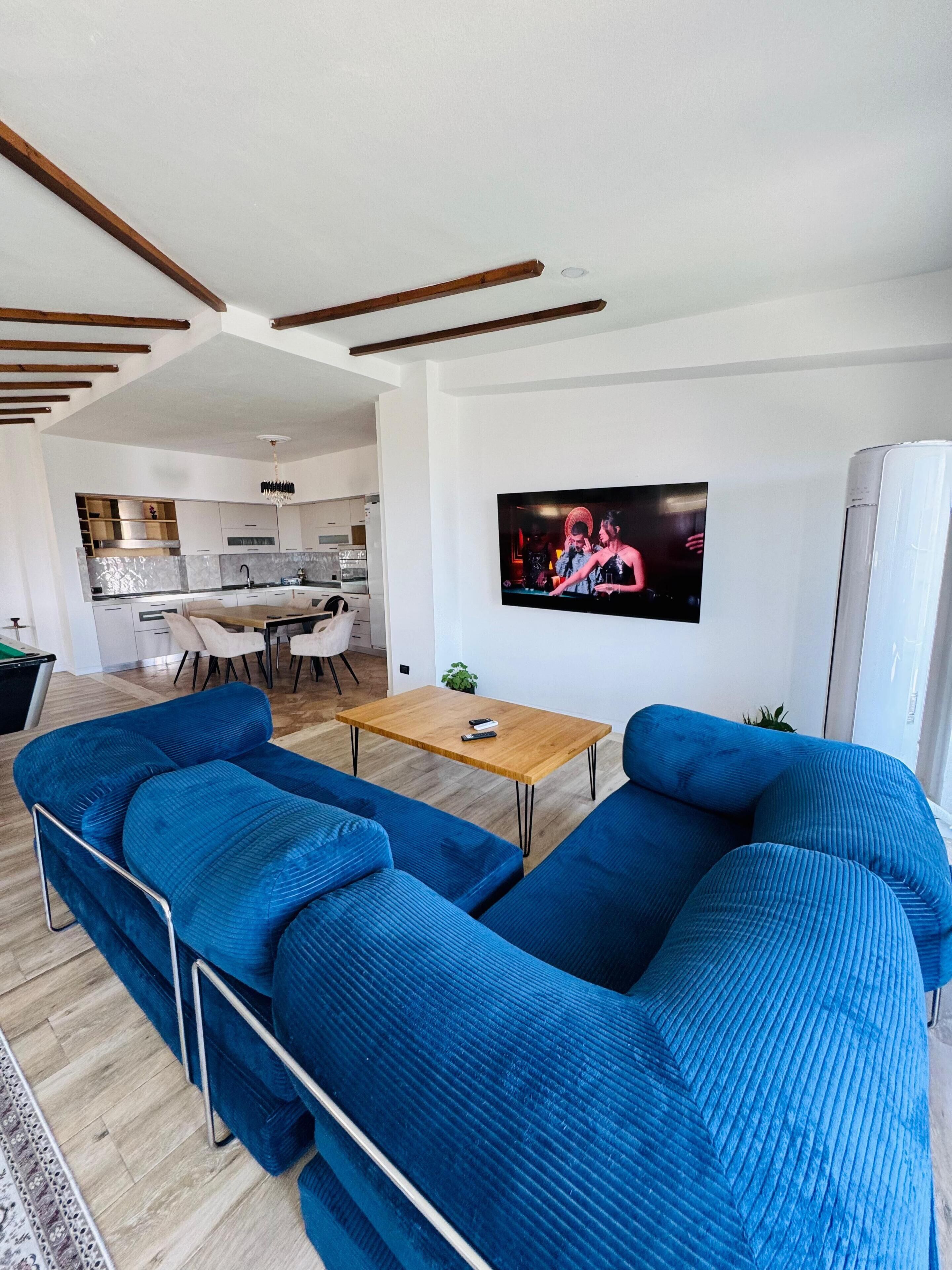 Een moderne woonkamer met een grote blauwe bank, een houten salontafel en een flatscreen televisie die aan de muur is bevestigd.