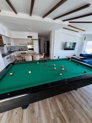 Ein Billardtisch mit Kugeln, eine Küche mit weißen Schränken, ein Essbereich mit Stühlen und ein Wohnzimmer mit einer Couch.