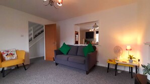 A living room with a sofa, a chair, a small table with a lamp, a fan, and a staircase. - Number One - A colourful family friendly space! (Crossways)