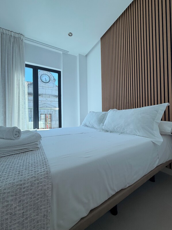 Habitación de hotel con cama, ventana con cortinas y vistas a un edificio, además de un panel de pared de madera.