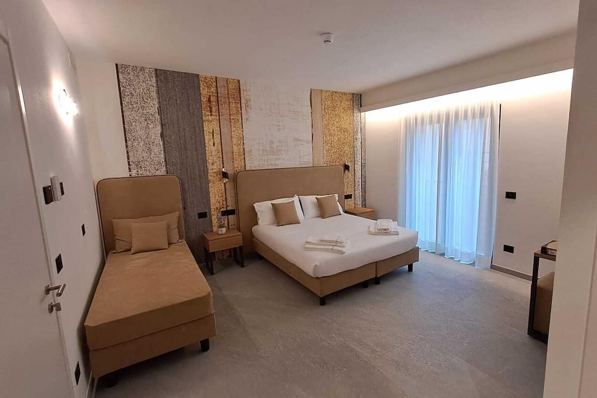 Una habitación de hotel con cama, sofá, mesita de noche y un ventanal con cortinas.