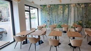 Moderni ruokailutila, jossa on puupöydät ja -tuolit, riippuvalaisimet ja seinämaalaus, jossa on kuvattuna puita.