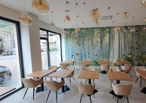 A modern dining area with wooden tables and chairs, hanging pendant lights, and a wall mural of trees. - MOUNTAIN SUITE - Hotel Apartment (Tirano)