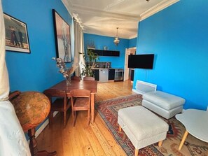 Un salon avec une table à manger en bois, un canapé, une petite télévision et, au fond, un coin cuisine.