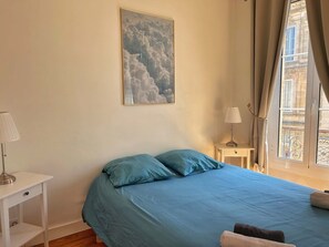Un dormitorio con cama, mesita de noche, lámpara, un cuadro y una ventana con cortinas.
