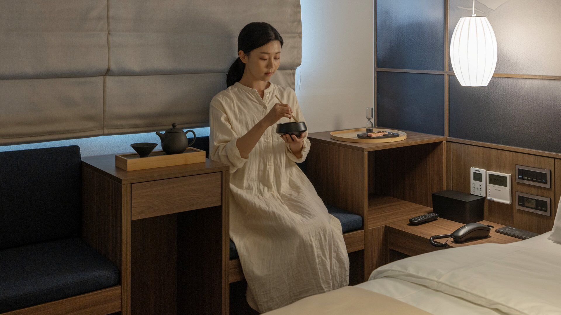 A hotel room with a bed, bedside tables, a desk with a tea set, a lamp, and a control panel.