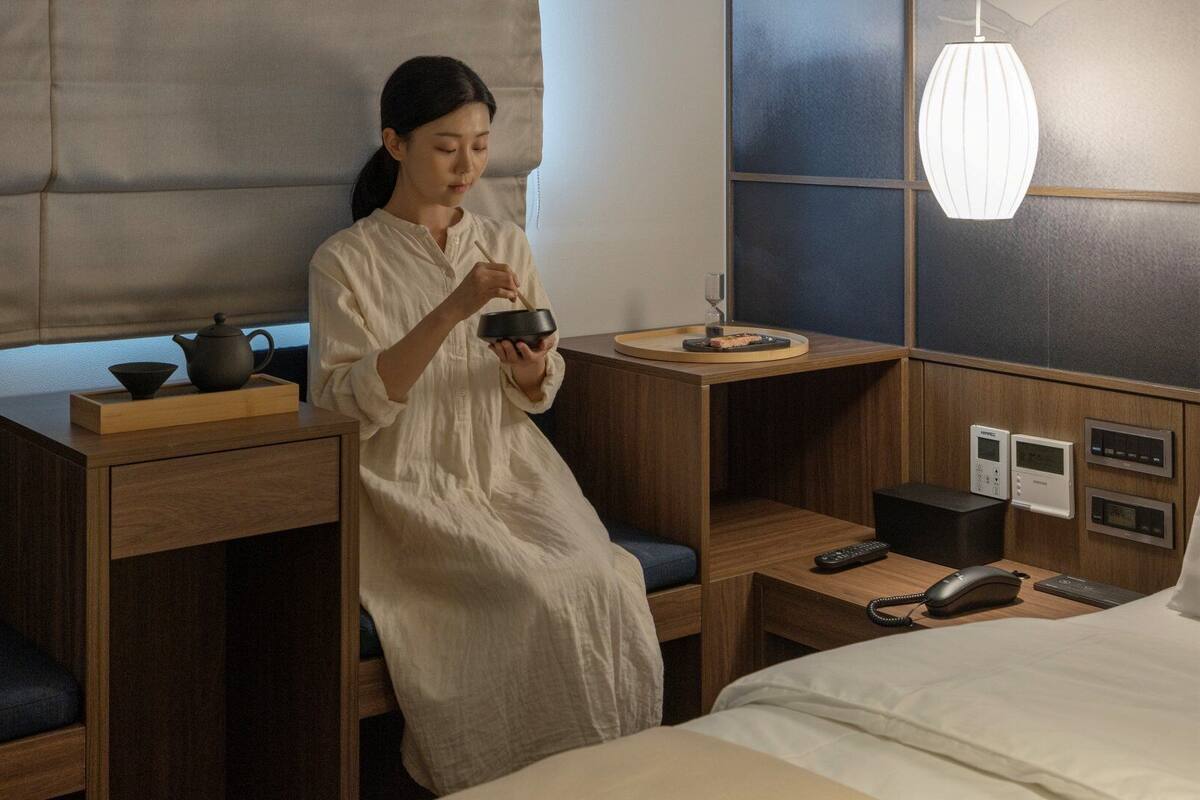 A hotel room with a bed, bedside tables, a desk with a tea set, a lamp, and a control panel.