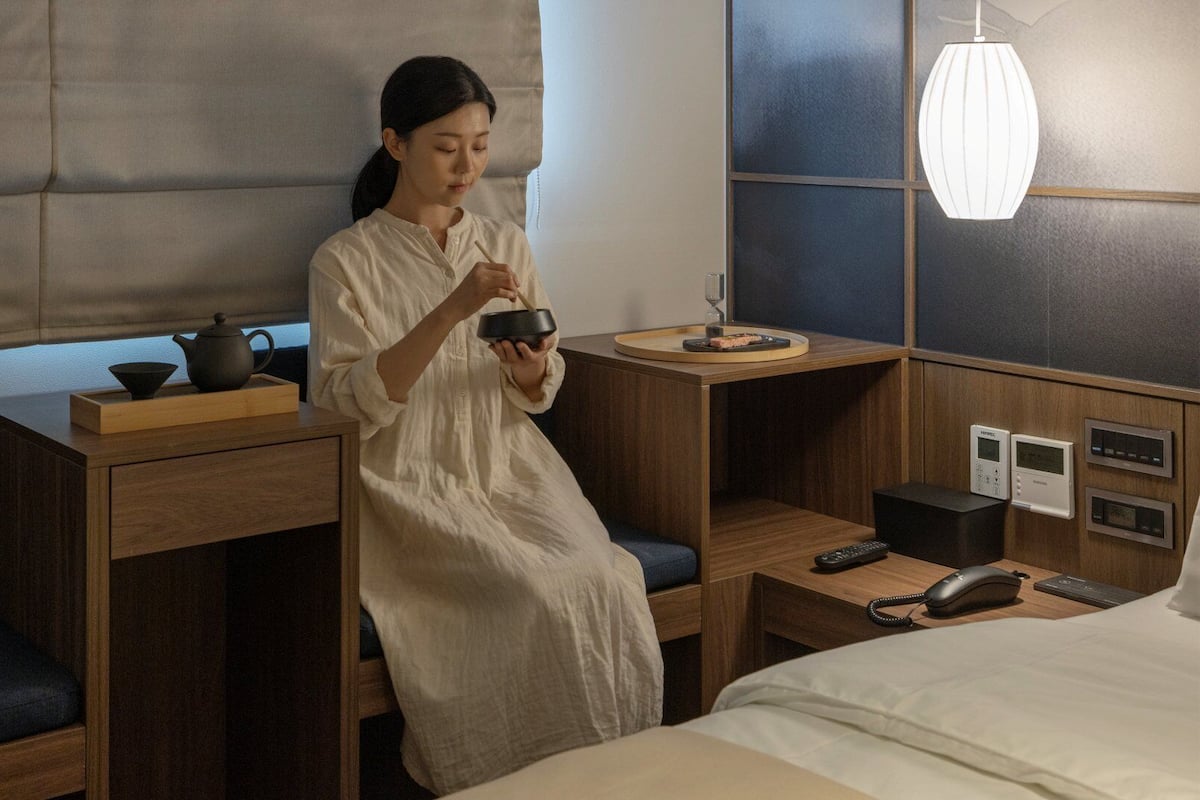 Una habitación de hotel con cama, mesitas de noche, un escritorio con juego de té, una lámpara y un panel de control.