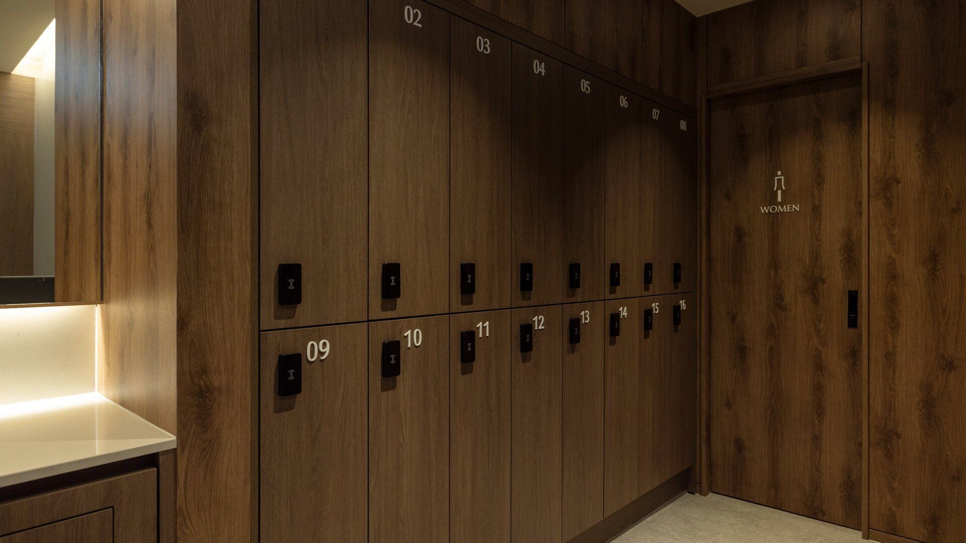 A row of wooden lockers with numbers 01 to 16, a women's sign on the right, and a lighted counter to the left.