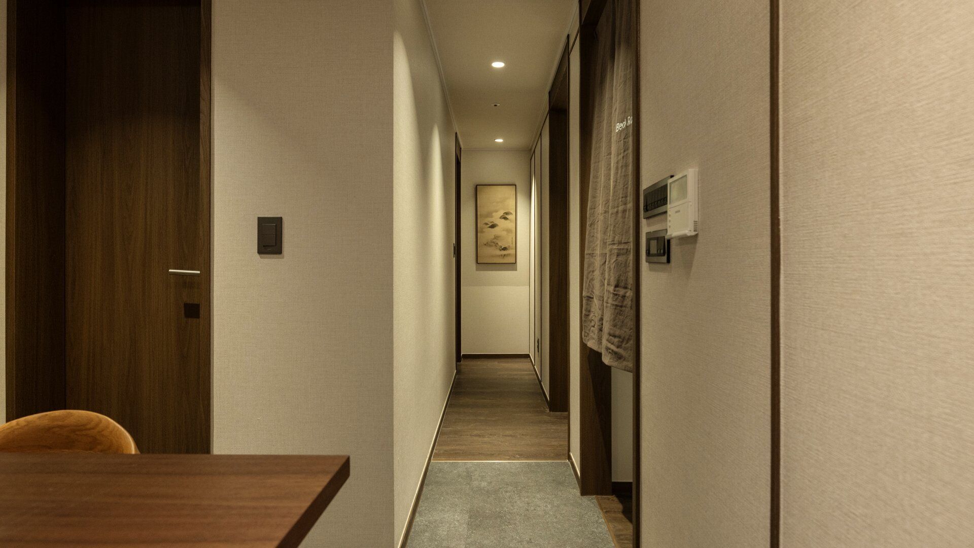 A narrow hotel corridor with a wooden door, a framed artwork on the wall, and a small table with a chair.