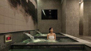 Une personne dans un bain à remous, avec de l’eau qui coule, une horloge numérique et un téléviseur fixé au mur.
