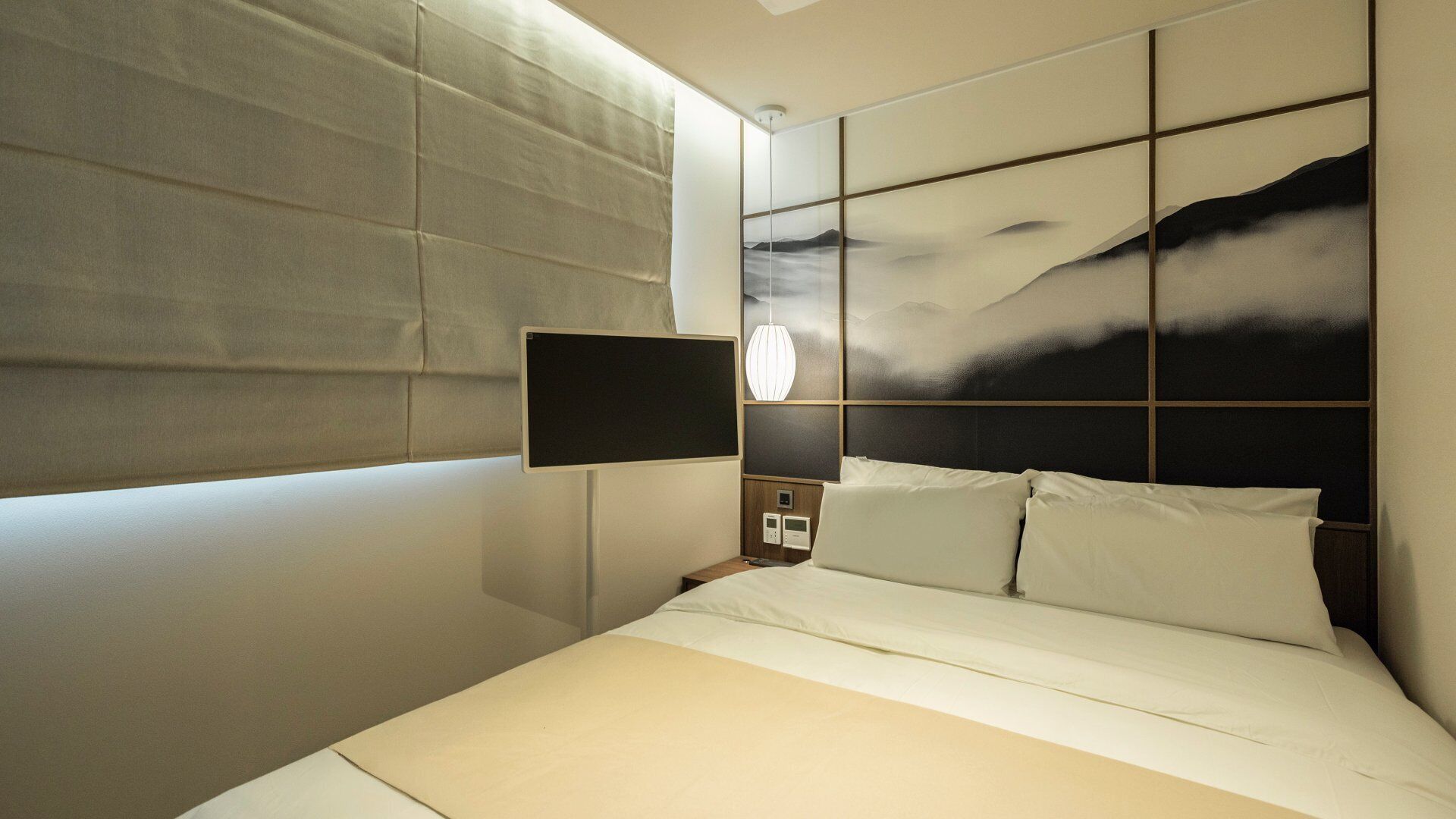 A modern hotel room with a large bed, a mounted TV, a bedside table with a lamp, and a view of mountains through the window.