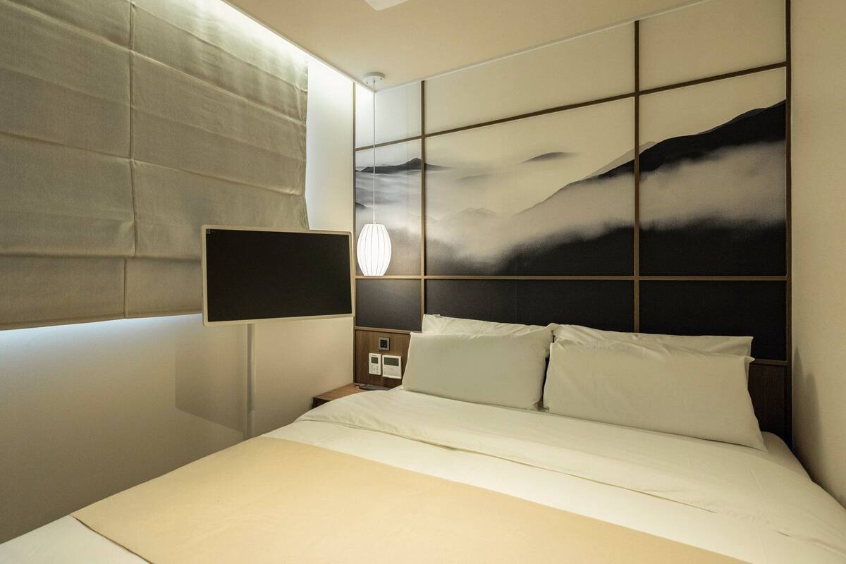 A modern hotel room with a large bed, a mounted TV, a bedside table with a lamp, and a view of mountains through the window.