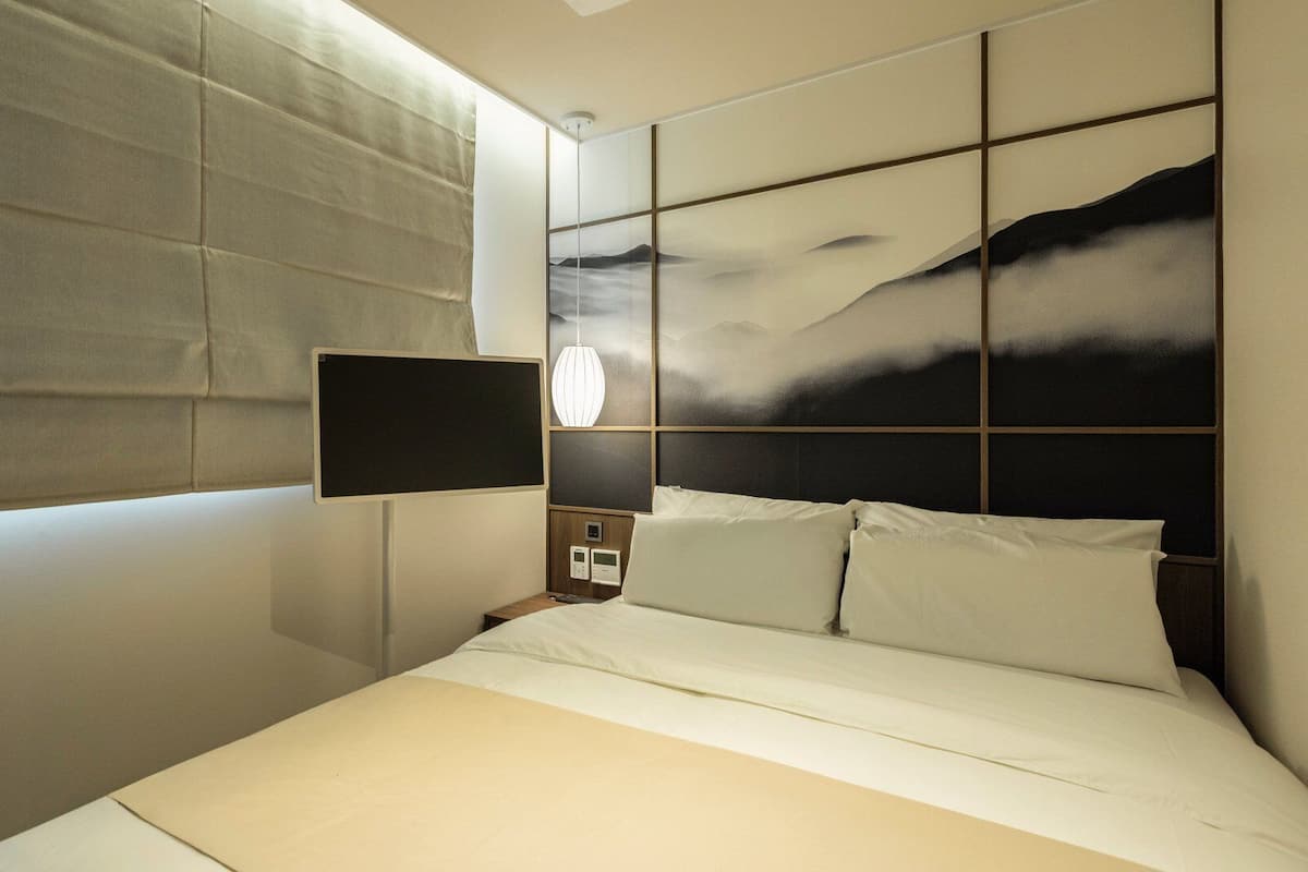 Un hotel moderno con una cama grande, un televisor montado, una mesita de noche con lámpara y vistas a las montañas a través de la ventana.