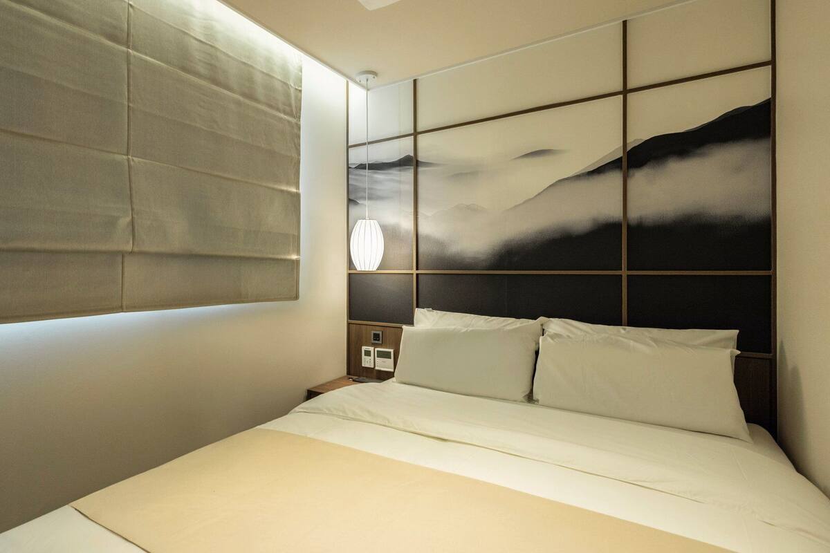 A modern hotel room with a large bed, a nightstand, a wall-mounted lamp, and a view of the mountains through the window.