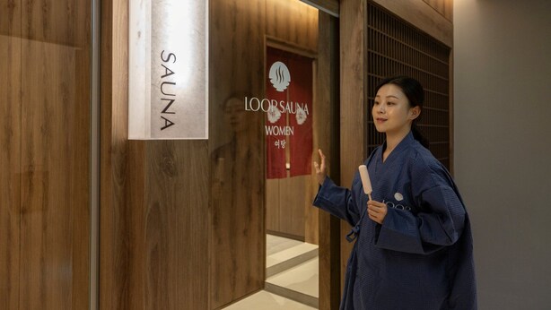 Une personne vêtue d’une robe bleue se tient dans un couloir, devant un panneau indiquant l’emplacement d’un sauna et un autre panneau indiquant l’emplacement d’un sauna réservé aux femmes.