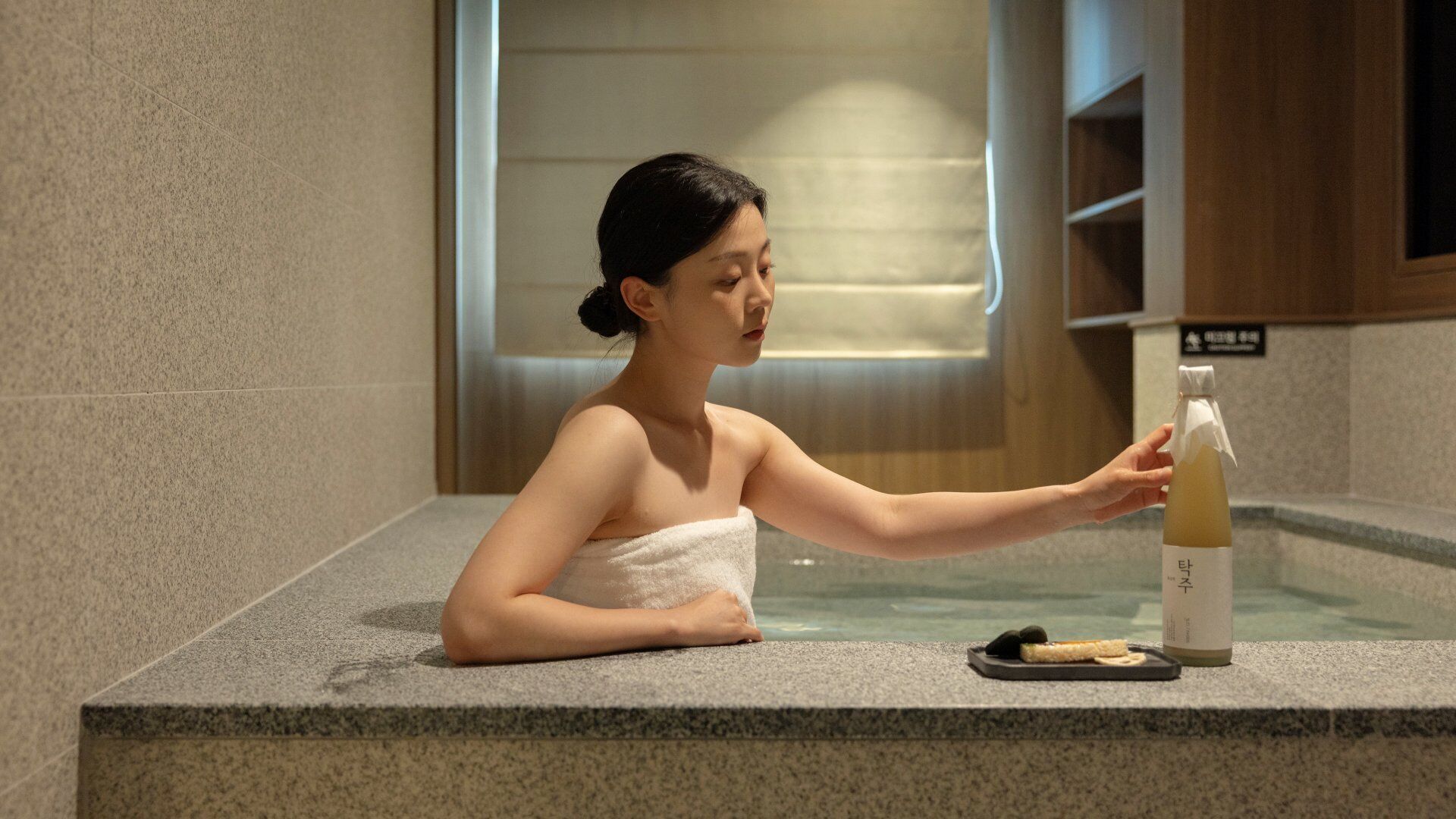A person in a spa setting with a towel wrapped around their body, a bottle of liquid, and a small plate with food items on a stone counter.