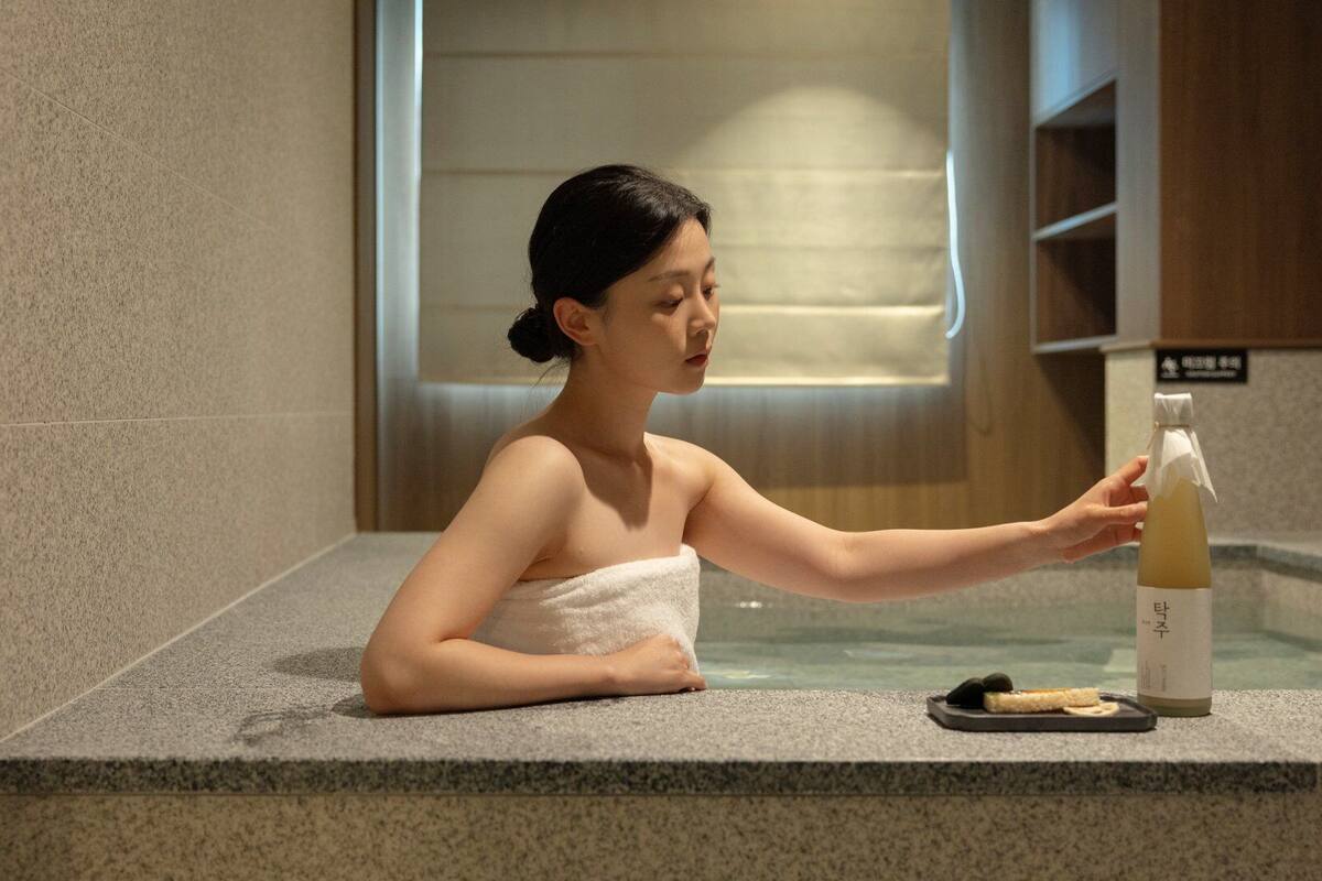 A person in a spa setting with a towel wrapped around their body, a bottle of liquid, and a small plate with food items on a stone counter.